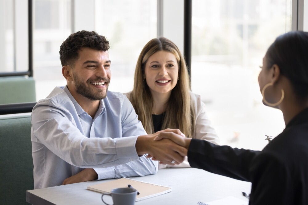 Cheerful young couple shaking hands with real estate agent, thanking for consultation.